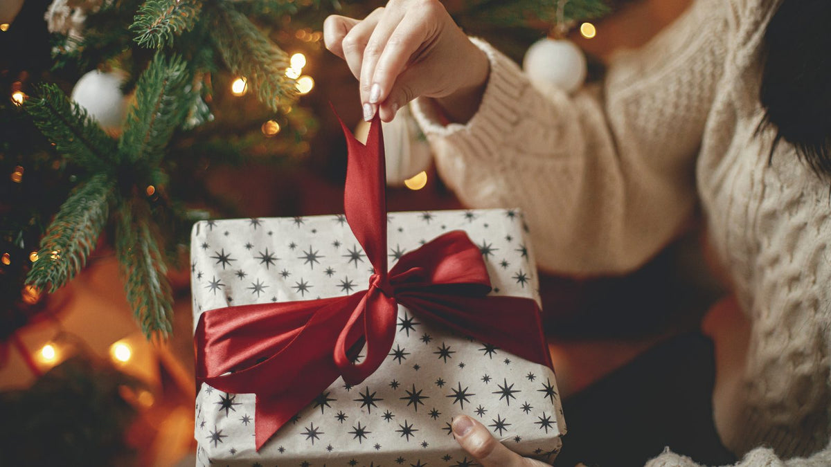 Mains qui dénouent le ruban rouge d'un cadeau de Noël emballé, au pied d'un sapin illuminé