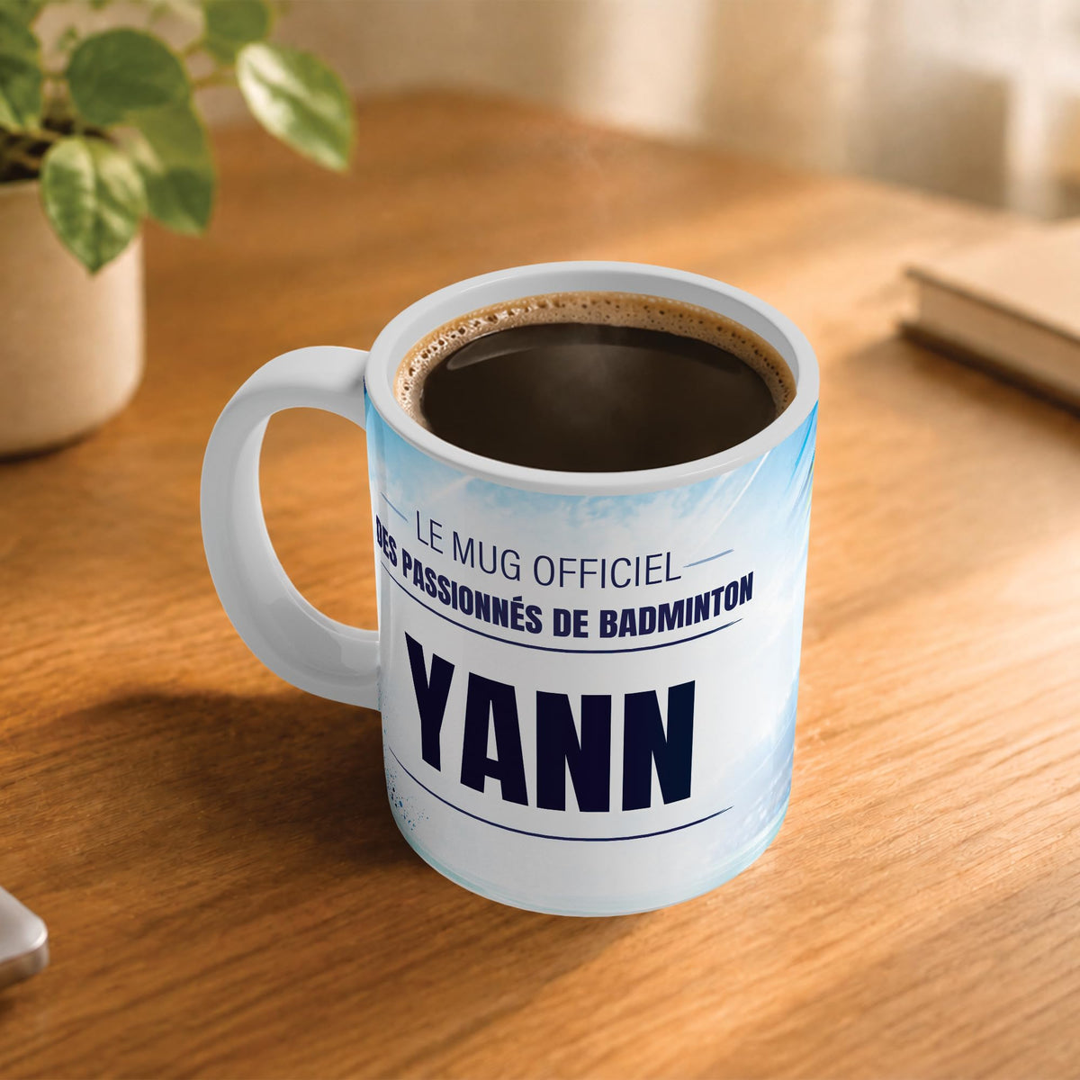 Café fumant dans le mug badminton posé sur un bureau en bois : la phrase « Le mug officiel des passionnés de badminton » et son prénom en grandes capitales, mug livré chez vous (exemple)